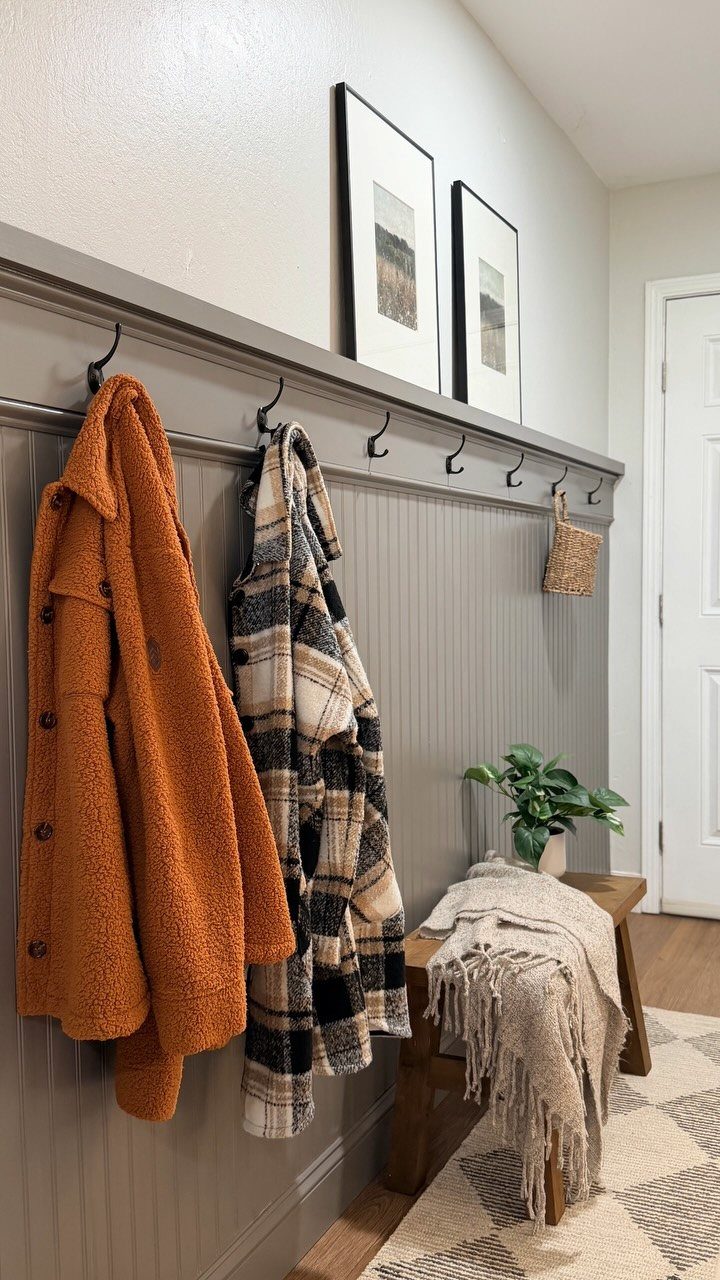 Grey beadboard accent wall with coat hooks, hanging jackets, and a wooden bench below.