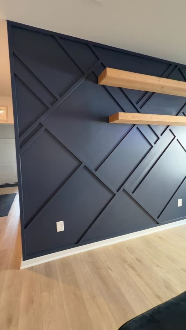 Navy blue geometric accent wall with two light wood floating shelves on a wooden floor.