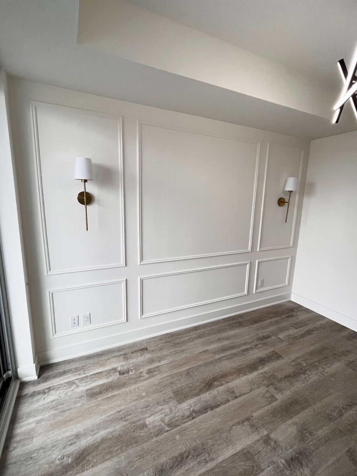 White wall with decorative molding and gold sconces in a room with grey flooring.