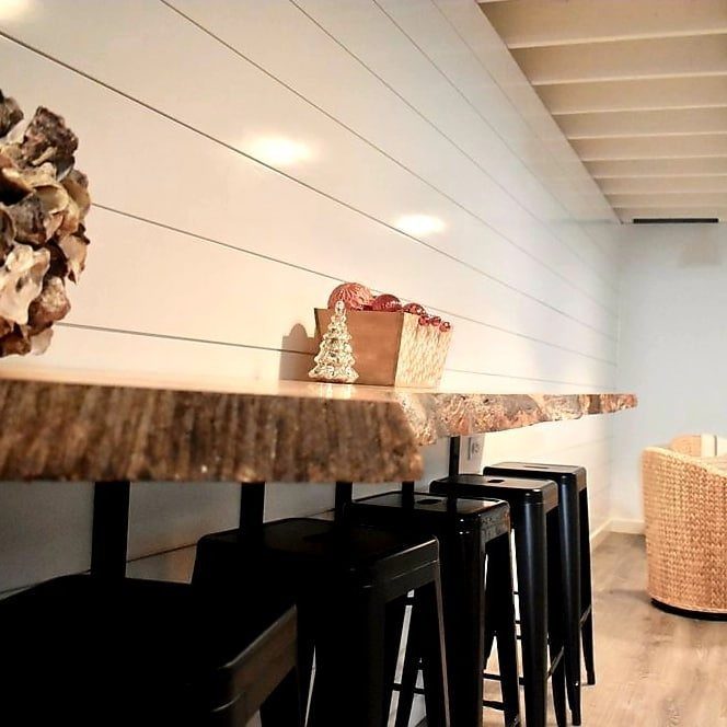 White shiplap accent wall behind a long live-edge wooden bar counter with black stools.