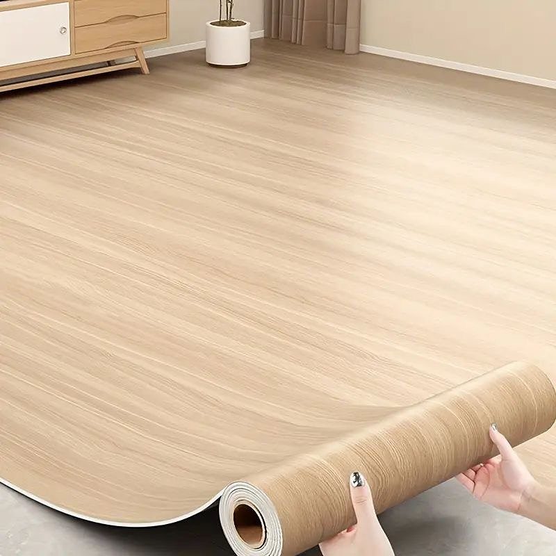 10 Affordable Flooring Ideas for Every Room in 2026 10 Hands unrolling a light wood-grain patterned sheet of vinyl flooring in a room.