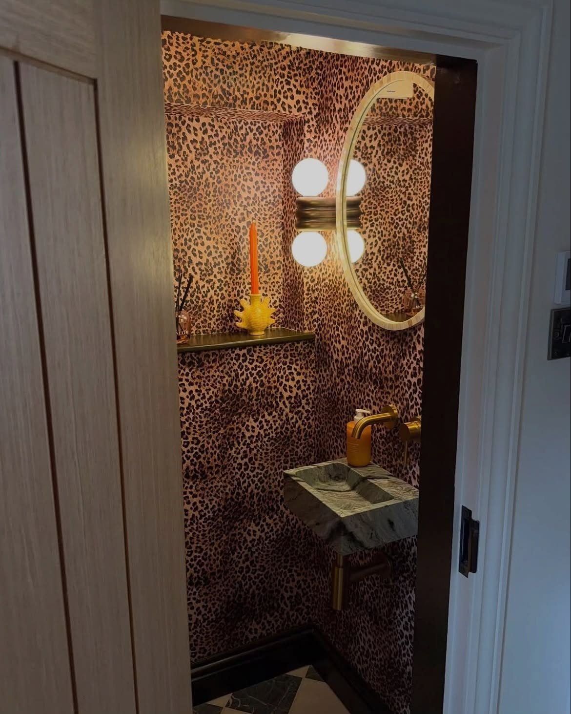 Small bathroom with leopard print wallpaper, marble sink, and round mirror with globe lights.
