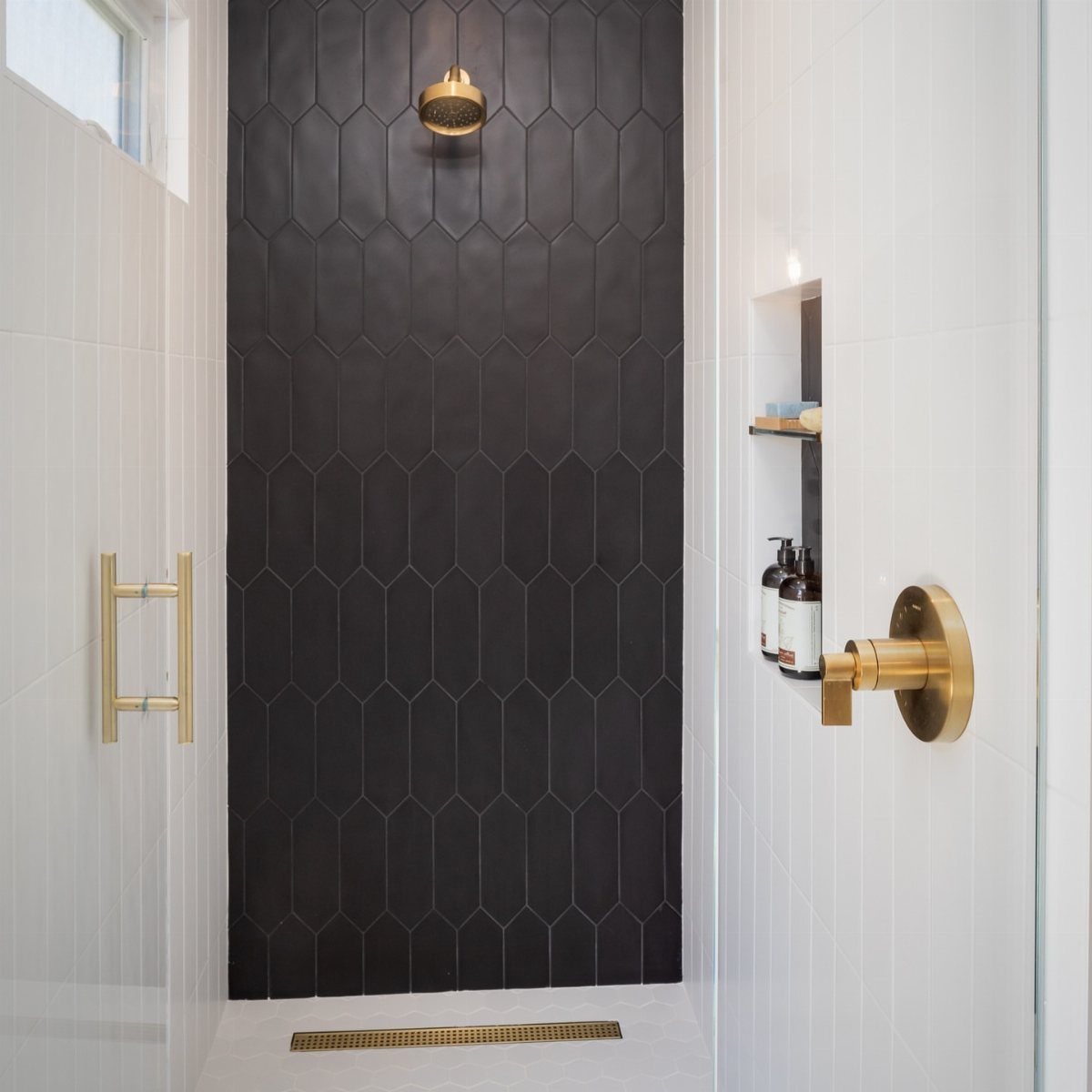 Black elongated hexagonal tiles form a dramatic accent wall inside a modern shower with gold fixtures.