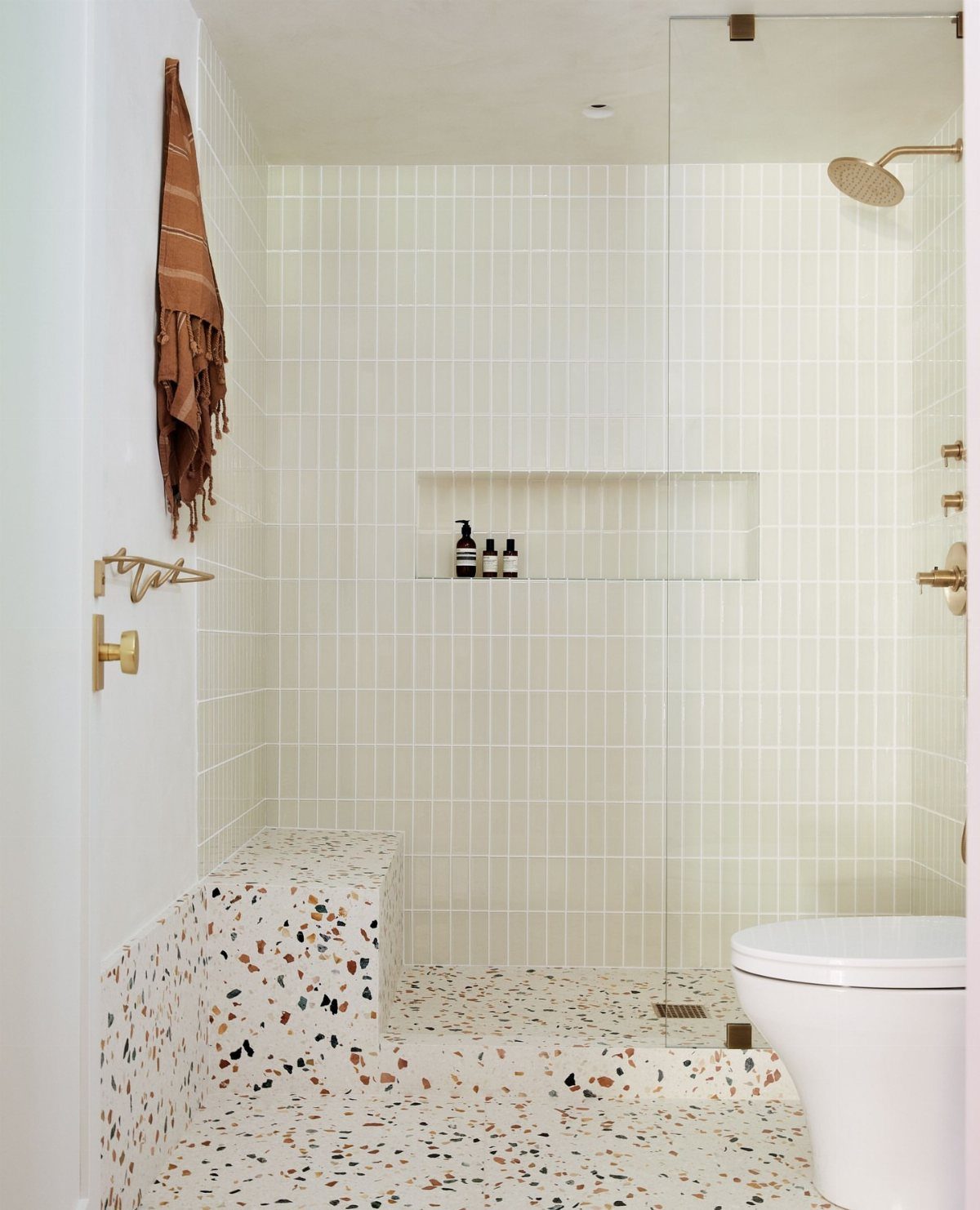 White vertical rectangular tiles on a bathroom wall with terrazzo flooring and gold fixtures.