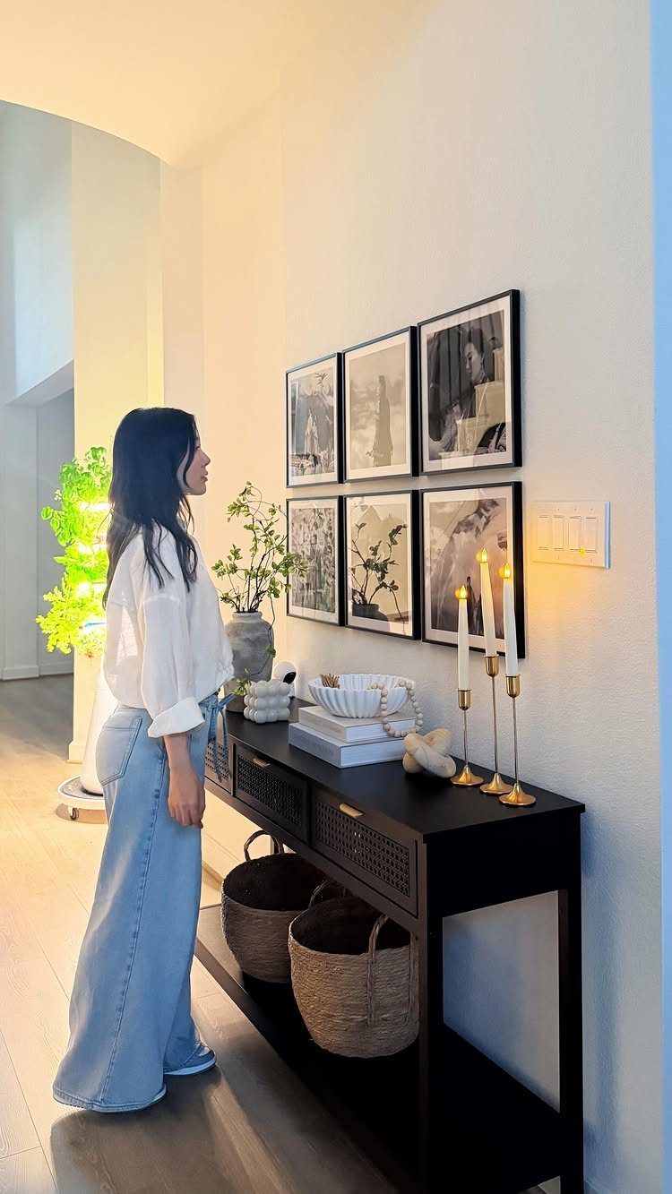 15 Feature Wall Ideas That Rely on Material, Not Just Color 4 A woman stands beside a black console table featuring a curated gallery wall of framed art.