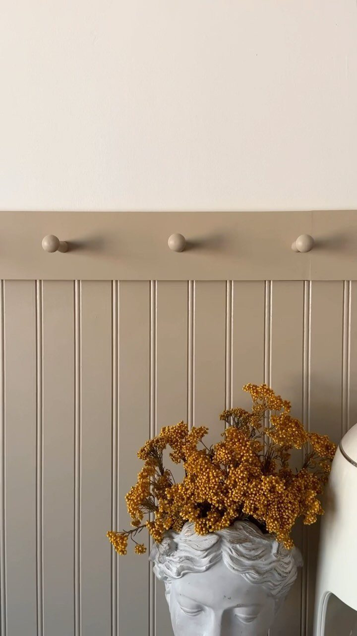 Beige beadboard wall with a chair rail and decorative pegs behind a white bust with yellow flowers.