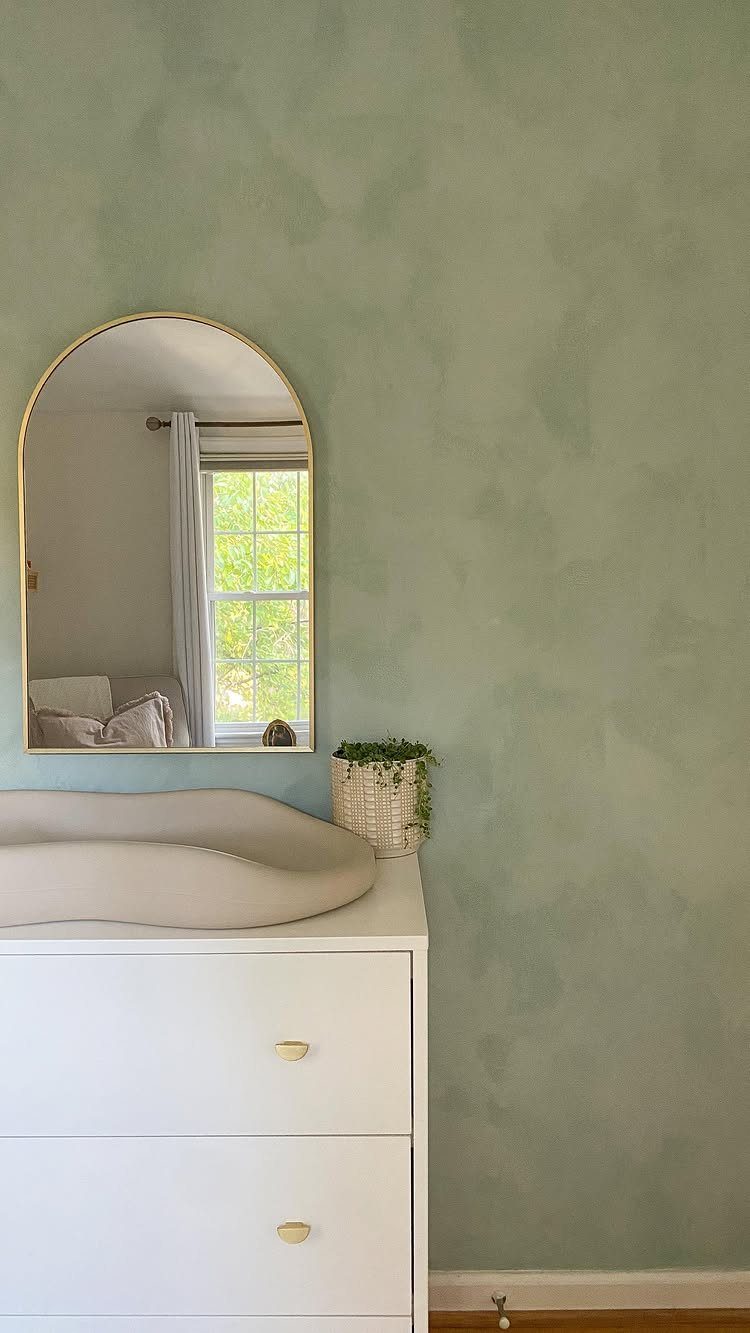 Sage green limewash textured wall behind a white dresser and an arched gold mirror.