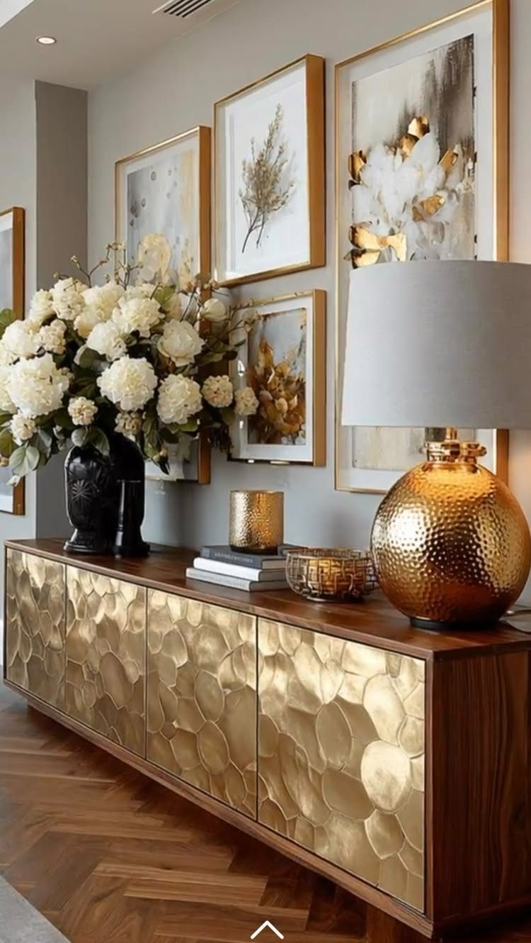 Gold-framed botanical art prints arranged on a grey wall above a wooden sideboard with flowers.