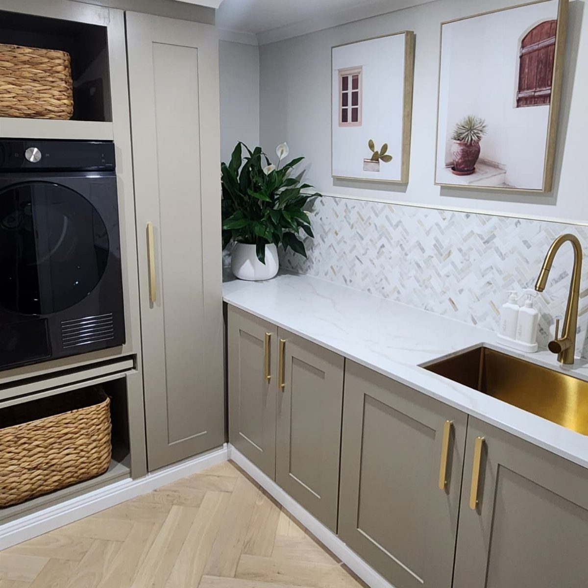 Light wood herringbone flooring in a modern laundry room with grey cabinets and gold accents.