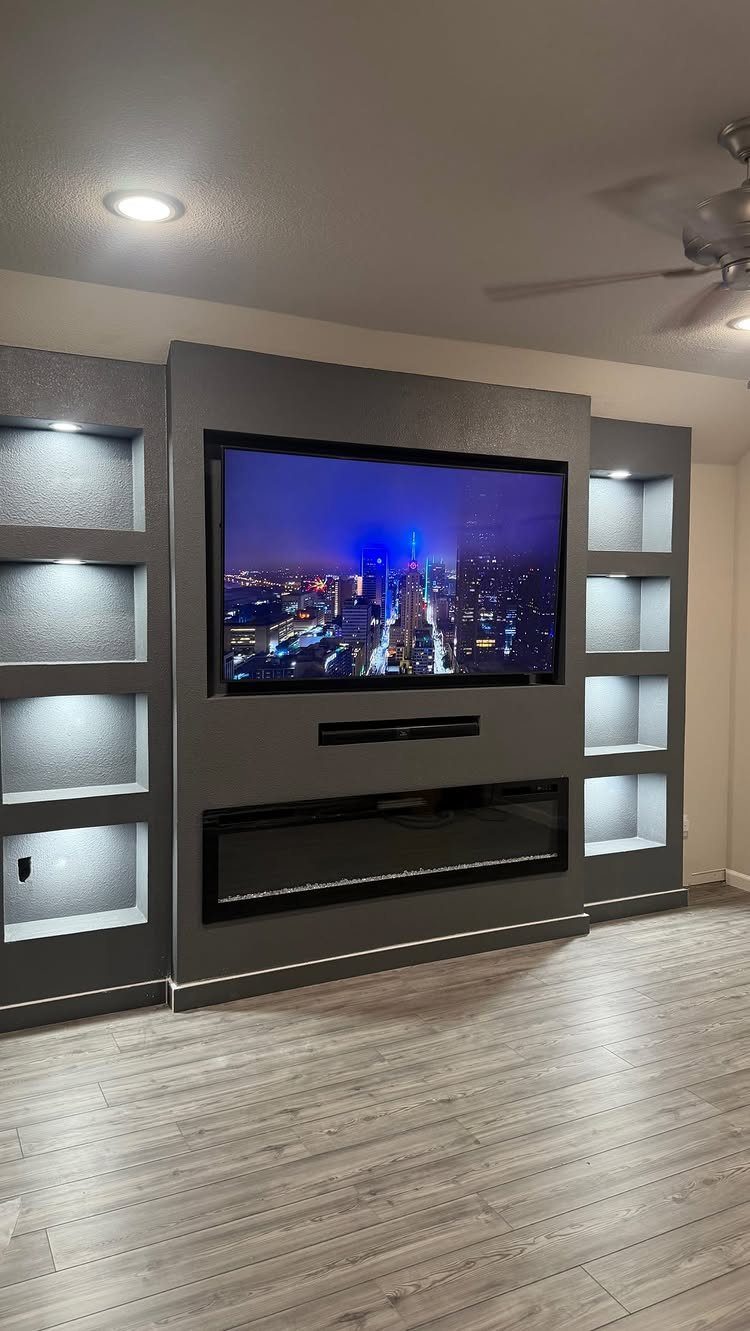 A gray recessed media wall with a television, soundbar, fireplace, and lit shelving units.