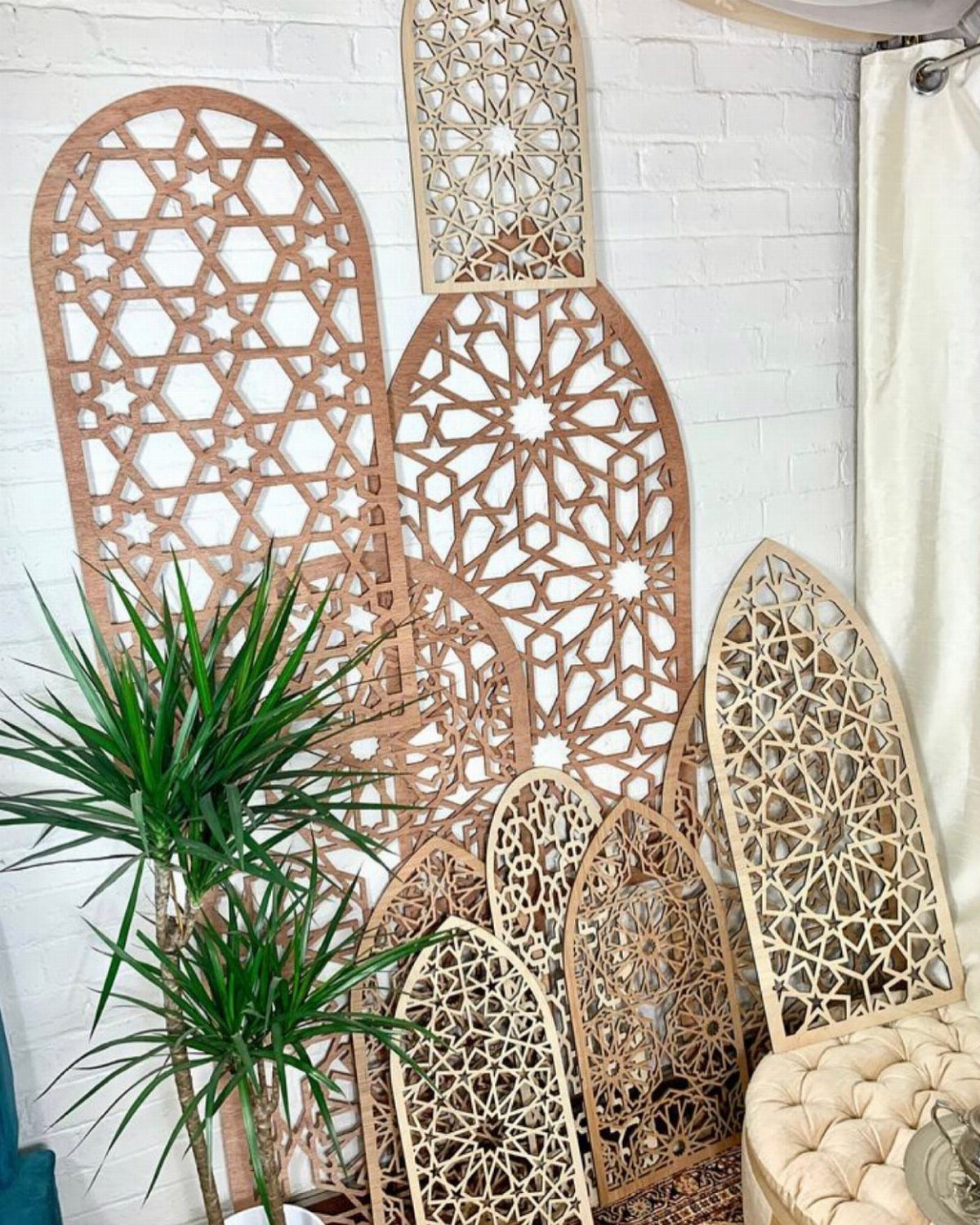 Intricate laser-cut wooden Moroccan-style geometric panels leaning against a white brick wall near a green plant.