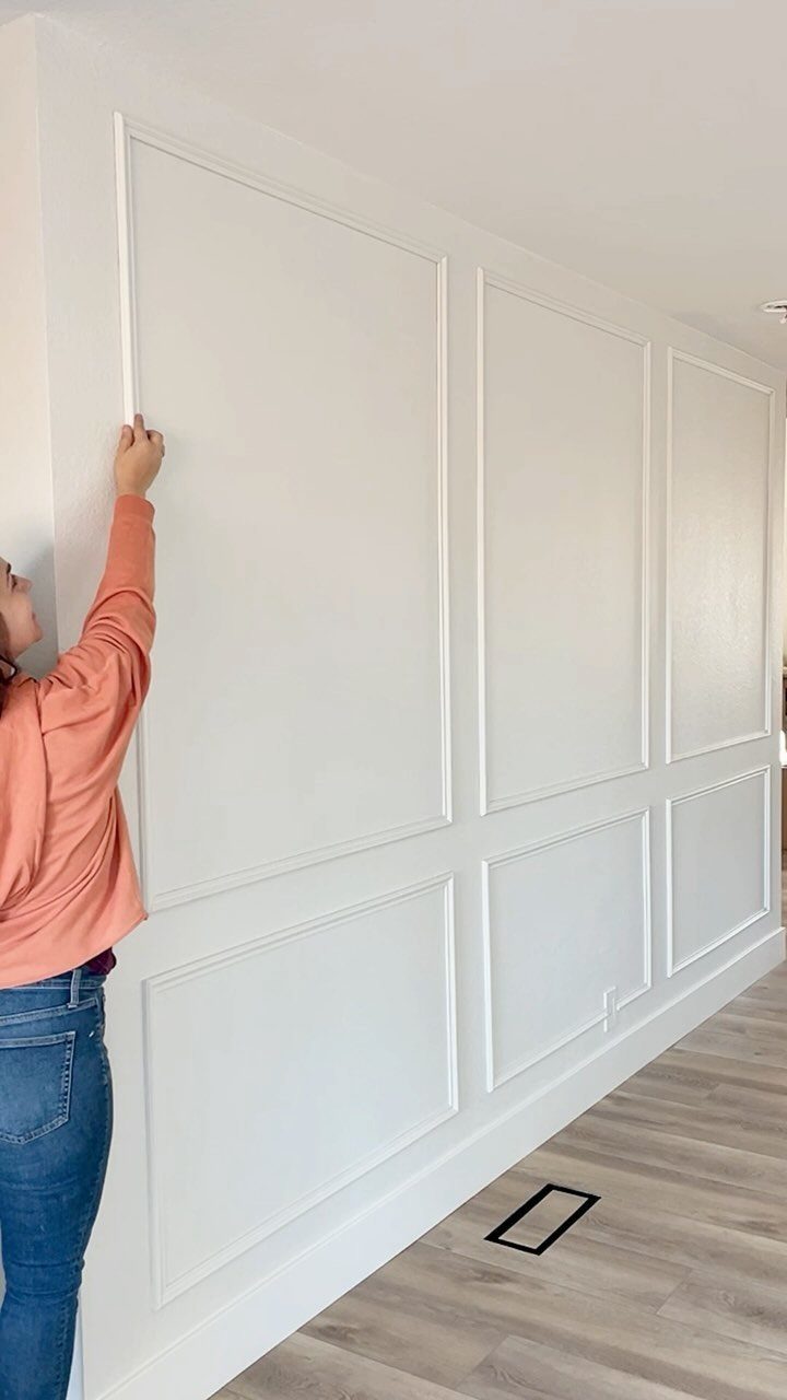 A person installing white decorative molding on a wall to create large rectangular panels.