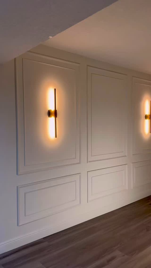 White wall with decorative rectangular panel molding and glowing gold vertical sconces mounted on the panels.