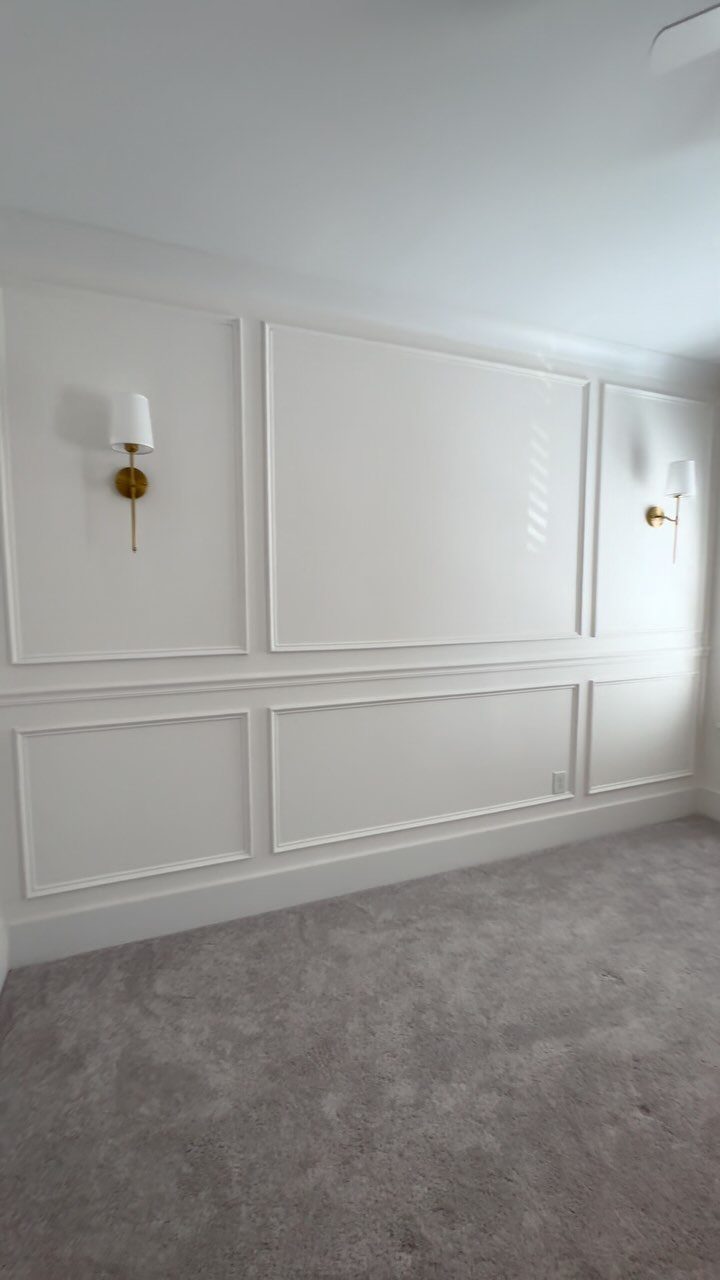 White wall with decorative rectangular panel molding and gold sconces above grey carpet.