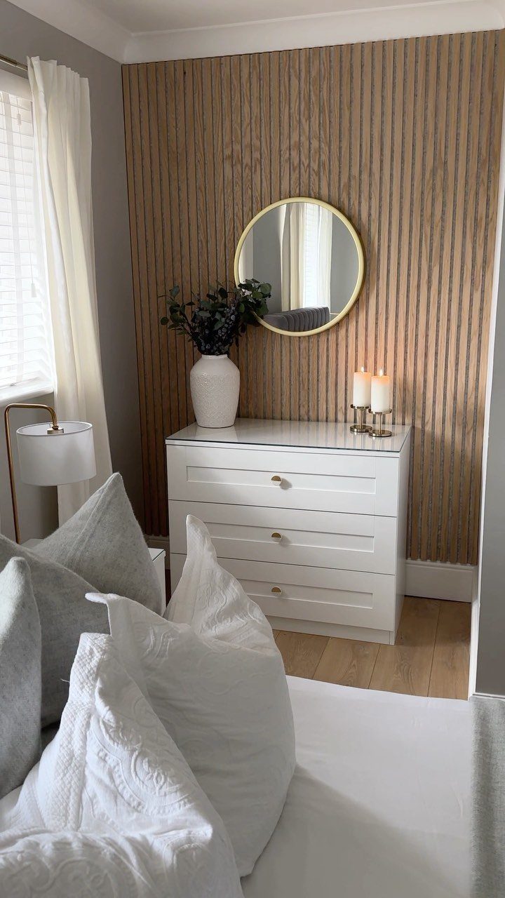 Vertical light wood slats cover a bedroom wall behind a white dresser and gold mirror.