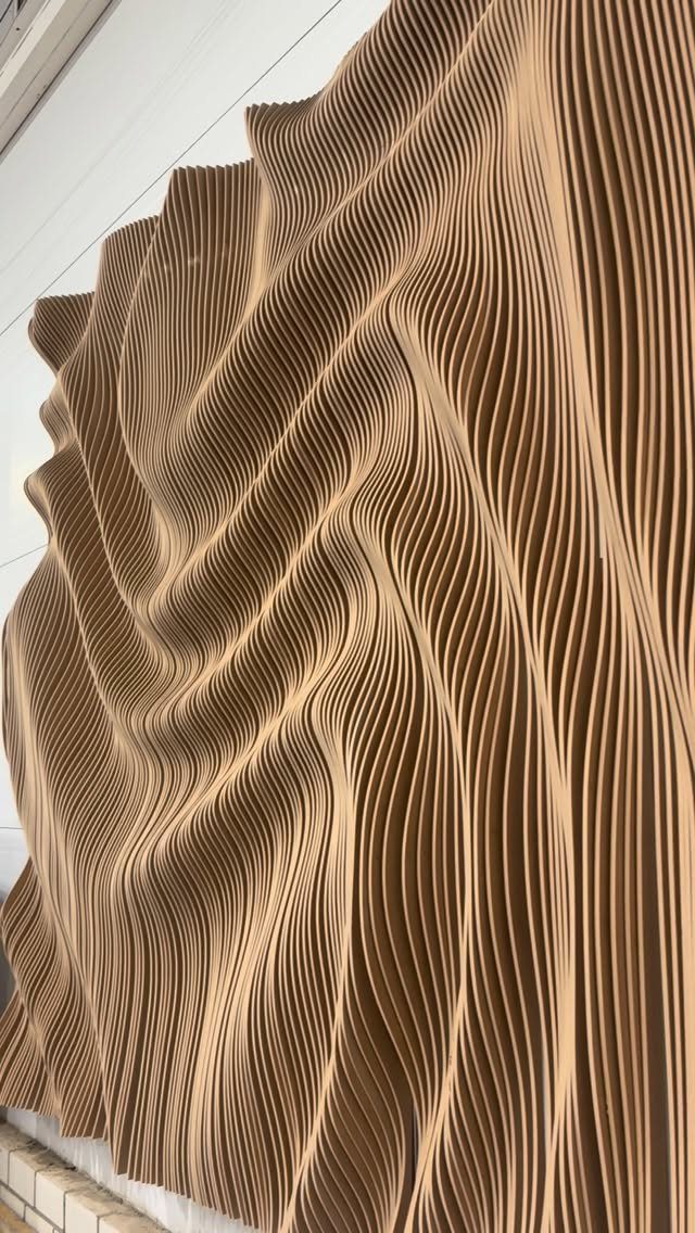 Undulating light brown wooden slats forming a large 3D textured wave pattern on a wall.
